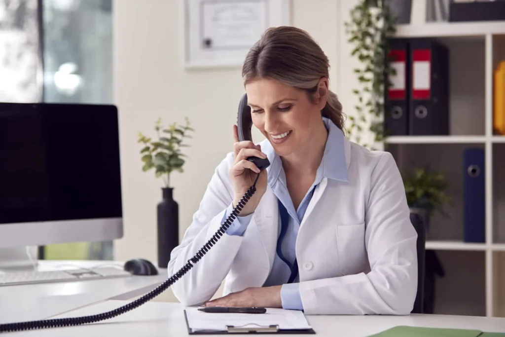 Smiling Female Doctor Or GP Wearing White Coat Sitting At Desk In Office Making Phone Call